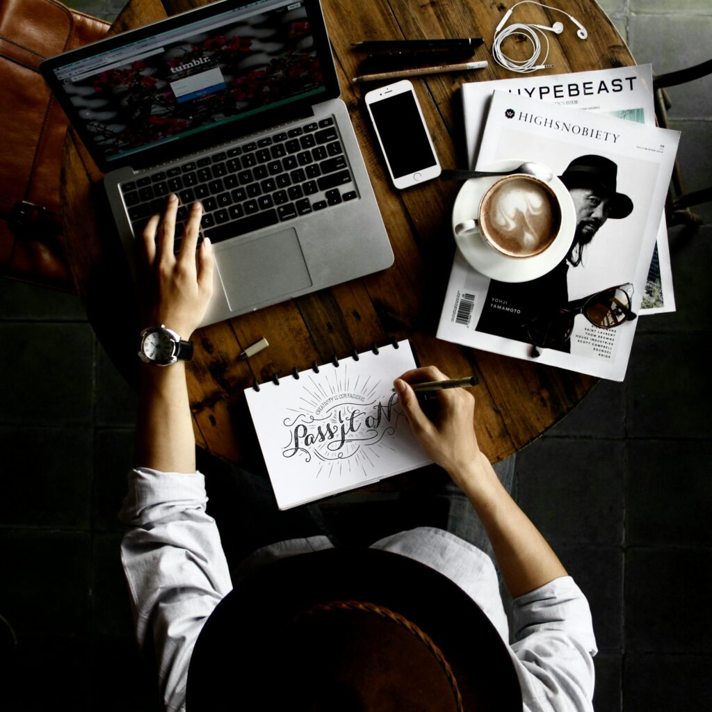 Top view of a creative workspace with a laptop, coffee, smartphone, magazines, and a notebook with hand lettering design.